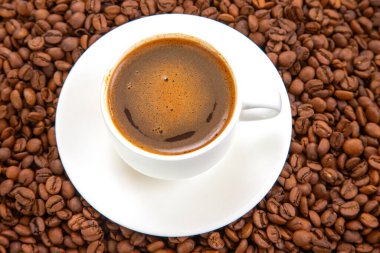 Beyaz seramik bir fincanda köpüklü krema ile zengin, koyu espresso. Dağılmış kahve çekirdekleri arasında zarif bir şekilde bir fincan üzerine yerleştirilip sıcak, davetkar bir kahve sahnesi yaratıyor.