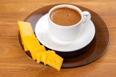 Beyaz bir fincanda köpüklü bir espresso, bir fincan üzerinde dinleniyor, sıcak ahşap bir masa yüzeyinde canlı sarı peynir dilimleri eşliğinde