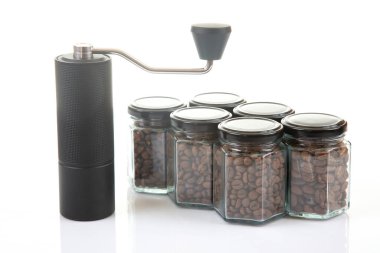 Kahve çekirdekleri için siyah manuel kahve öğütücüsü ve siyah kahve çekirdekleri ile dolu altı bardak şeffaf kavanoz ve beyaz arka planda kahve demlemek için toprak takımı..
