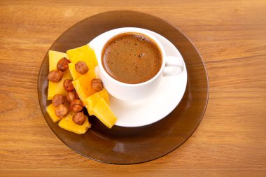 Beyaz bir fincanda köpüklü bir espresso, bir fincan üzerinde dinleniyor, sıcak ahşap bir masa yüzeyinde canlı sarı peynir dilimleri eşliğinde