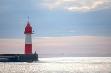 Kızıl ve beyaz deniz feneri, sakin bir denize ve sakin bir sabah boyunca yumuşak, bulutlu bir gökyüzüne karşı yalnız bir balıkçıyla bir iskelede dimdik ayakta duruyor..