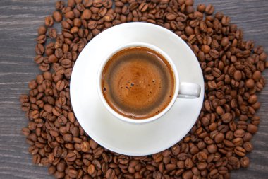 Klasik bir beyaz espresso fincanı ve fincan tabağı, kavrulmuş kahve çekirdekleri ile çerçevelenmiş, zengin, köpüklü bir kahve tutar.