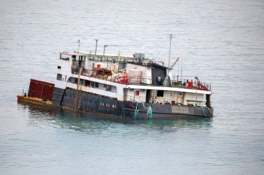 Sakin deniz sularında yüzen paslı eski kargo gemisi soluk boya ve dağınık güverte ile aşınma izleri gösteriyor. Geminin kırmızı ve beyaz gövdesi bulutlu bir gökyüzünün altındaki dingin mavi okyanusla çelişiyor..