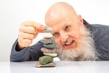 Hevesli, yaşlı bir adam, çeşitli taşlar yığını inşa ederken geniş bir gülümseme içinde, sükuneti, sabrı ve taş istifleme yoluyla doğayla bağlantı kurmanın sakinleştirici uygulamasını sembolize ediyor.