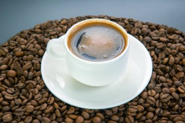 Beyaz bir fincandaki dumanı tüten bir espressonun üst görüntüsü, altın kreması, etrafındaki kavrulmuş kahve çekirdekleri ile tezat oluşturuyor, hepsi de beyaz bir tabağın üzerinde duruyor..