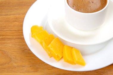 Tabağın üzerinde köpüklü bir yüzeye sahip beyaz bir fincan espresso 'nun yukarıdan görünüşü, ahşap bir masanın üzerinde sarı peynir dilimleriyle eşleştirilmiş. Minimalist ve kırsal ayar.