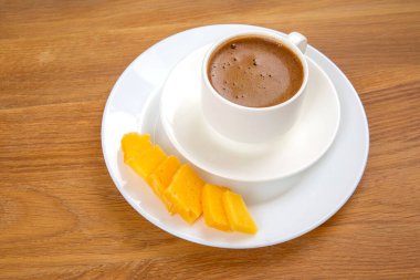 Zengin espressoyla dolu beyaz bir fincanın yukarıdan aşağı görüntüsü, köpüklü bir üst, sıcak ahşap masadaki sarı peynir dilimlerinin yanına konmuş bir fincan..