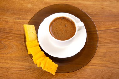 Zengin espressoyla dolu beyaz bir fincanın yukarıdan aşağı görüntüsü, köpüklü bir üst, sıcak ahşap masadaki sarı peynir dilimlerinin yanına konmuş bir fincan..