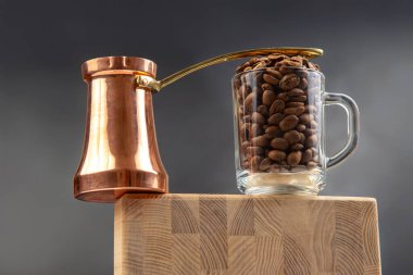 Geleneksel bir bakır çakmak ve kavrulmuş kahve çekirdekleri ile dolu bir cam bardak, koyu mavi arka planda, ahşap bir yüzeye oturur. Kırsal ve zarif bir kahve temalı kompozisyon..