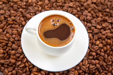 Koyu kahve ve kremalı beyaz espresso bardağının yukarıdan görünüşü, uyumlu bir fincan tabağına yerleştirilmiş, kahverengi desenli kahve çekirdekleri ile çevrili.