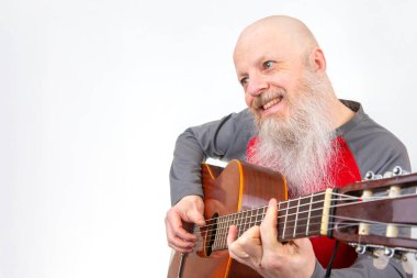 Saçsız ve uzun beyaz sakallı yaşlı bir adam gitar çalıyor, kırmızı ve gri atletik bir bluz giyiyor, beyaz bir arka planda fotoğrafı çekiliyor, bir müzik hobisini vurguluyor.