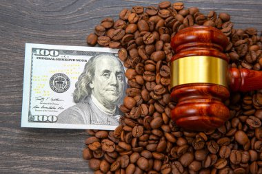 Tahta bir tokmak, koyu ahşap bir yüzey üzerinde 100 dolarlık bir banknotun yanında duran kahve çekirdekleri, hukuk, finans ve ticaretin kesişimini sembolize ediyor. Yasal, ticari veya ticari temalar için ideal
