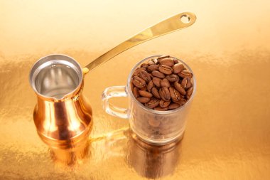 Pirinç saplı bakır bir kahve demliği ve yansıtıcı arka planda kahve çekirdekleri ile dolu bir cam bardak. Sıcak, zarif bir ses tonuyla mutfak, kültür veya lüks mutfak eşyaları fotoğrafçılığı için idealdir.