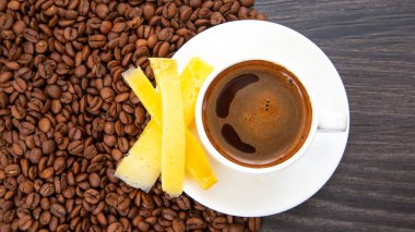 Beyaz bardakta dumanı tüten bir espressonun üst görüntüsü, yanında sarı peynir çubukları ve koyu ahşap arka planda dağınık kahve çekirdekleri var..