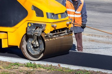 Bir inşaat sahasında siyah asfaltı sıkıştıran sarı bir yol pateninin yakın çekimi. Makinenin büyük varili ve tekerleği görünür, yan tarafında çakıllar ve çimenler var..