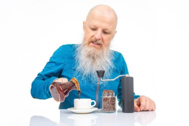 Beyaz sakallı ve kel kafalı son sınıf öğrencisi kahve öğütücüsü kullanıyor, kahve çekirdeği kavanozu ve seramik bir kafenin yanında, parlak beyaz bir yüzey üzerinde..