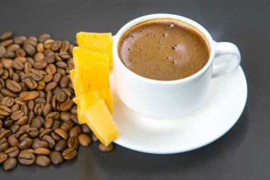Kavrulmuş kahve çekirdekleri koyu bir yüzeyde beyaz bir fincan espresso ve sarı peynir çubuklarını sarıp sıcak ve davetkar bir kompozisyon oluşturur..