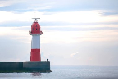 Sessiz bir iskelede, kırmızı ve beyaz bir fenerde, sakin bir okyanus ve sakin bir gökyüzü ile çevrili, sahilin huzurlu özünü yakalıyor..