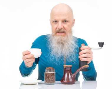 Uzun beyaz sakallı kel yaşlı bir adam, kahve çekirdeklerini öğütücüyle öğütüyor, elinde beyaz bir fincan, bir kavanoz fasulye ve yansıtıcı beyaz bir masa üzerinde geleneksel bir merhem tutuyor..