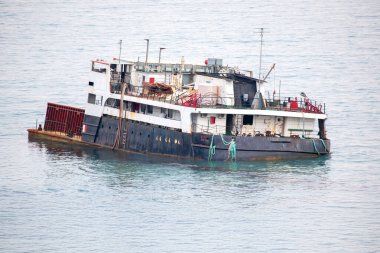 Sakin deniz sularında yüzen paslı eski kargo gemisi soluk boya ve dağınık güverte ile aşınma izleri gösteriyor. Geminin kırmızı ve beyaz gövdesi bulutlu bir gökyüzünün altındaki dingin mavi okyanusla çelişiyor..