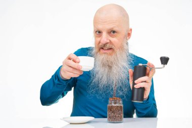 Uzun beyaz sakallı kel yaşlı bir adam, kahve çekirdeklerini öğütücüyle öğütüyor, elinde beyaz bir fincan, bir kavanoz fasulye ve yansıtıcı beyaz bir masa üzerinde geleneksel bir merhem tutuyor..