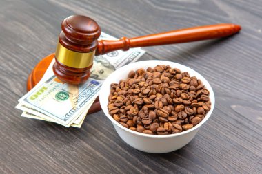 Gavel ve 100 dolarlık banknotlar, bir kase kavrulmuş kahve çekirdeğiyle ahşap üzerinde, yasal işlemlerin ve kahve ticaretinin temaları gösteriliyor..
