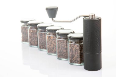 Kahve çekirdekleri için siyah manuel kahve öğütücüsü ve siyah kahve çekirdekleri ile dolu altı bardak şeffaf kavanoz ve beyaz arka planda kahve demlemek için toprak takımı..