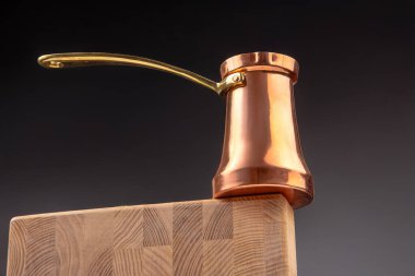 Pirinç saplı parlak bir bakır Türk kahve demliği, koyu renk bir arka plan, kahve kültürü ve mutfak teması için ideal olan ahşap bir kenarda dengede duruyor.