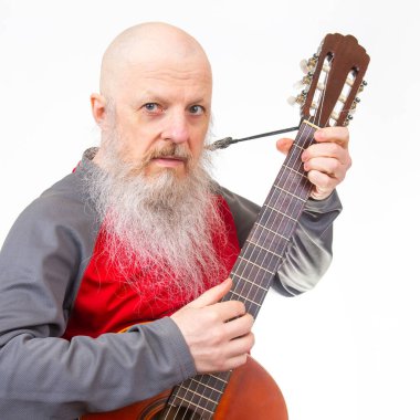 Uzun beyaz sakallı kel yaşlı bir adam akustik gitar çalar, kırmızı ve gri atletik bir gömlek giyer, beyaz bir arka planda izole edilmiş, bir müzik anı yakalar..