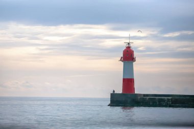 İskelede kırmızı ve beyaz bir deniz feneri olan sakin bir sahil sahnesi, bir balıkçı oltasını atıyor, ve şafak vakti yumuşak bir pastel gökyüzünün altında sakin bir deniz..