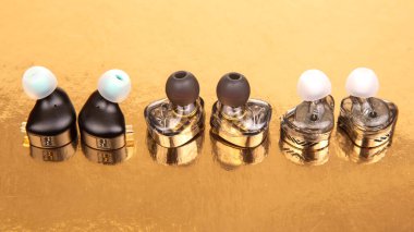 Yansıtıcı altın bir yüzey üzerinde siyah, gümüş ve şeffaf desenli üç çift parlak kulaklık. Teknoloji, ses ve yaşam tarzı temaları için ideal. Yüksek çözünürlüklü fotoğraf.