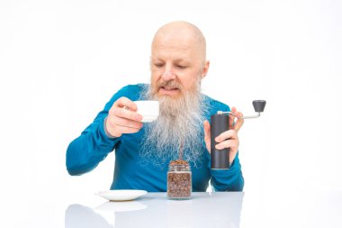 Beyaz sakallı ve kel kafalı son sınıf öğrencisi kahve öğütücüsü kullanıyor, kahve çekirdeği kavanozu ve seramik bir kafenin yanında, parlak beyaz bir yüzey üzerinde..