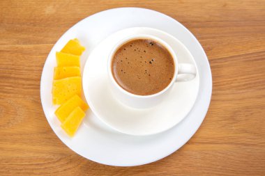 Tabağın üzerinde köpüklü bir yüzeye sahip beyaz bir fincan espresso 'nun yukarıdan görünüşü, ahşap bir masanın üzerinde sarı peynir dilimleriyle eşleştirilmiş. Minimalist ve kırsal ayar.