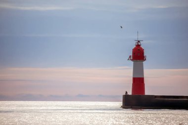 Kızıl ve beyaz deniz feneri, sakin bir denize ve sakin bir sabah boyunca yumuşak, bulutlu bir gökyüzüne karşı yalnız bir balıkçıyla bir iskelede dimdik ayakta duruyor..
