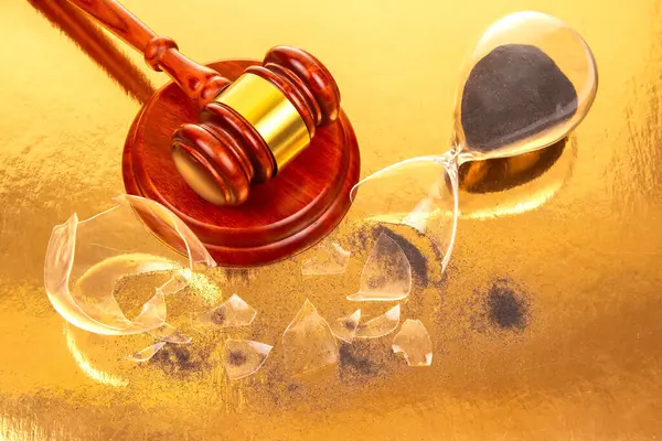 İyi işlenmiş ahşap bir tokmak, siyah kumlu kırık bir kum saatinin yanında duruyor, parıldayan altın bir zemin üzerinde fotoğraflanmış, hukuk ve zamansallık temalarını harmanlıyor.