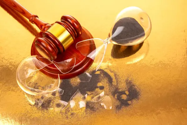 Altın bantlı ahşap bir tokmak, siyah kumla parçalanmış kum saatinin yanına yerleştirilir. Yansıtıcı altın bir yüzey üzerinde, hukuki kararların geçici doğasını gösteriyor..