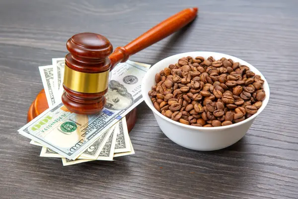 Adalet ve ticaret çarpışıyor: 100 dolarlık banknotların üzerinde bir tokmak ve yanında bir kâse kahve çekirdeği, ahşap bir masaya kurulmuş, hukuk ve ekonominin altını çiziyor..