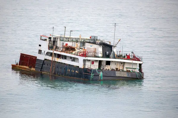 Sakin deniz sularında yüzen paslı eski kargo gemisi soluk boya ve dağınık güverte ile aşınma izleri gösteriyor. Geminin kırmızı ve beyaz gövdesi bulutlu bir gökyüzünün altındaki dingin mavi okyanusla çelişiyor..