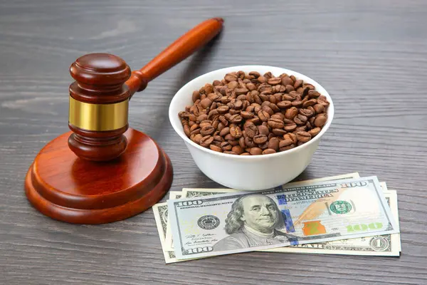 Tahta bir masada, 100 dolarlık banknotlar üzerinde bir tokmak ve bir kase kahve çekirdeği adalet, finans hukuku ve küresel kahve ticareti temalarını çağrıştırıyor..