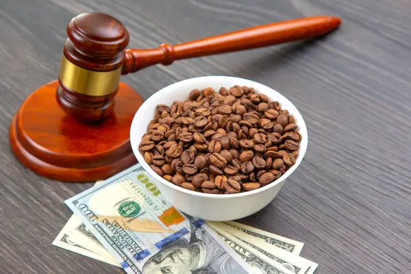 Hukuk ve ticareti sembolize eden tokmak, siyah ahşap bir masada beyaz bir kâse kahve çekirdeğinin yanında, adalet ve iş kavramlarını harmanlayan 100 dolarlık banknotlar üzerine kuruludur..