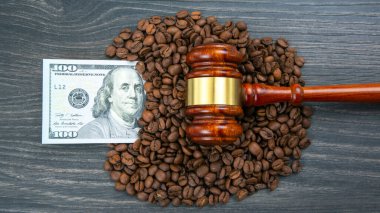 Kahve çekirdekleri ahşap bir tokmak ve koyu ahşap arka planda 100 dolarlık bir banknotla çevrili. Kahve ticareti ekonomisini ve ticaretin hukuki işleyişini simgeliyor.