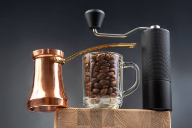 Parlak bakır bir kahve seti, bir bardak kavrulmuş kahve çekirdeği, ve ince bir manuel öğütücü, ahşap üzerine koyu arkaplan ile zarif bir şekilde düzenlenmiş.