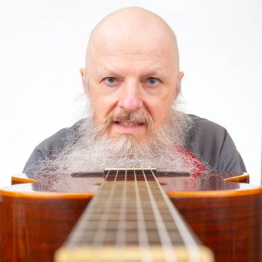 Uzun beyaz sakallı, kel, yaşlı bir adam akustik bir gitar tutuyor, gri atletik bir gömlek giyiyor, gitarı beyaz bir arka plana odaklıyor, bir müzik anı yakalıyor.
