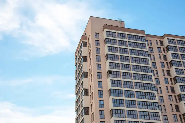 Açık mavi gökyüzünün altında bej bir cephesi ve büyük cam pencereleri olan modern bir gökdelen. Mimari tasarım temiz hatlara ve birden fazla balkona sahiptir.