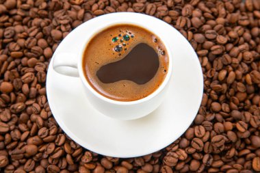 Koyu kahve ve hafif kremayla dolu beyaz bir espresso fincanı, tepeden tırnağa kapalı bir perspektiften yakalanmış, kavrulmuş kahve çekirdekleri ile çevrili, uyumlu bir fincan üzerinde oturur.