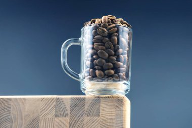 Kavrulmuş kahve çekirdekleri ile dolu şeffaf bir cam bardak koyu mavi arka planda, ahşap bir bloğun kenarında oturur. Minimalist bir natürmort kahve konsepti.