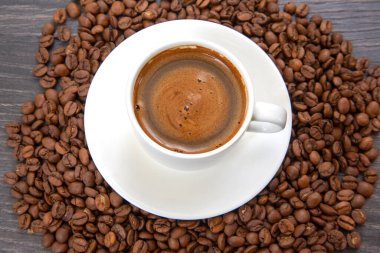 Yukardan bakıldığında kremalı köpüklü küçük beyaz bir fincan espresso sergileniyor. Bir fincan çay tabağının üzerinde ve koyu kavrulmuş kahve çekirdekleri ile çevrelenmiş..