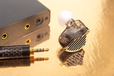 Bir kulaklık, 3.5 mm ses sistemi ve altın bir yüzeyde siyah bir ses cihazı. Teknoloji aksesuarlarını ve ses ekipmanlarını sergilemek için birebir..