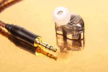 Beyaz uçlu tek bir kulaklık, 3.5 mm 'lik bir ses kablosu ve yansıtıcı altın bir yüzeyde siyah bir ses cihazı. Teknoloji ve ses temaları için ideal.