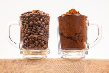 Ahşap bir yüzeyde iki temiz cam bardak, biri kavrulmuş kahve çekirdekleri, diğeri de ince öğütülmüş kahve tozu ile dolu. Kahve severler için mükemmel, işlenmemiş ve işlenmiş kahve sergileniyor..